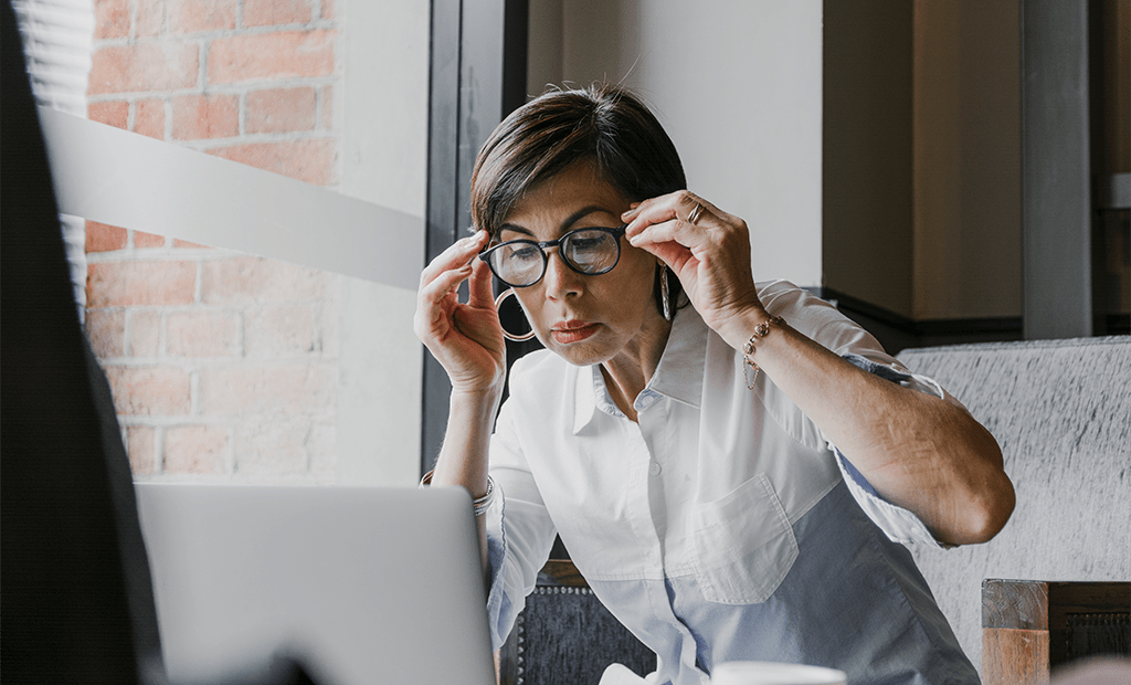 Autismo. Mulher de óculos observando atentamente a tela de um notebook, sentada em ambiente com parede de tijolos.