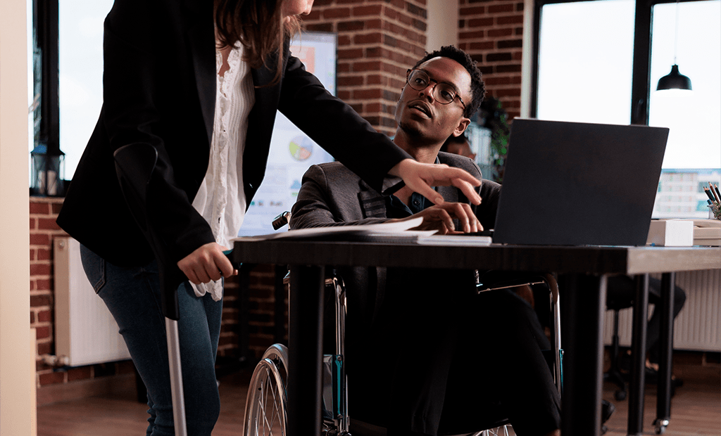 Overlays. Homem em cadeira de rodas trabalhando em um notebook, enquanto uma mulher com muletas ao lado aponta para algo na mesa. A cena acontece em um ambiente de escritório.