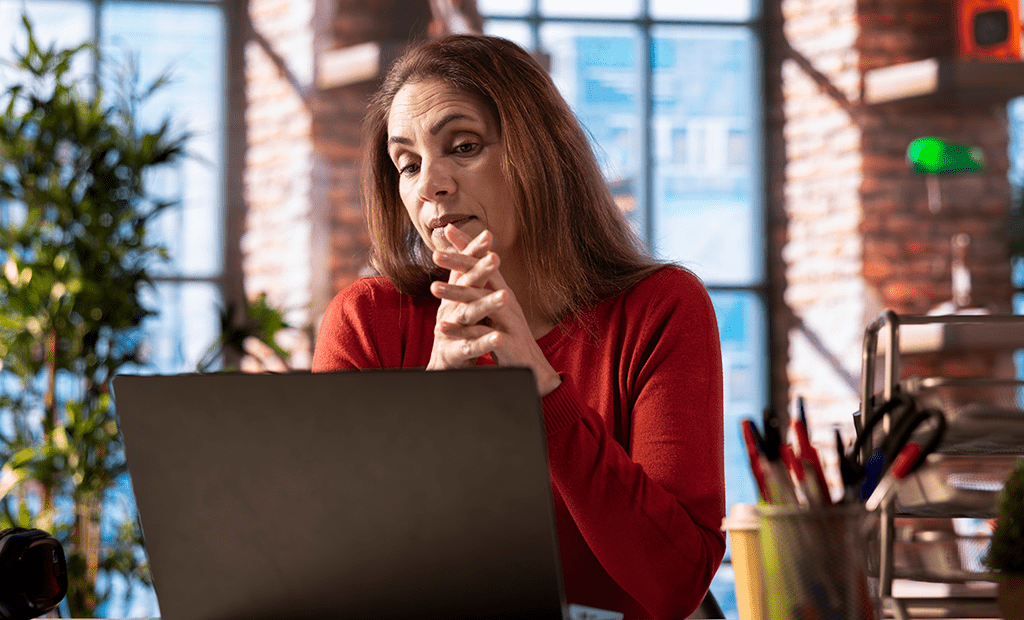 Acessibilidade digital no marketing. Mulher de blusa vermelha sentada em frente ao notebook, com expressão séria e mãos entrelaçadas, em um escritório iluminado.