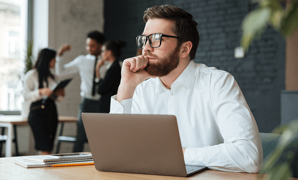 Acessibilidade digital no marketing. Homem de óculos e camisa branca observa pensativo enquanto trabalha no notebook, com colegas ao fundo conversando em um escritório moderno.