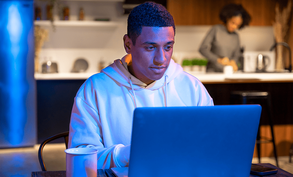 Acessibilidade digital no marketing. Homem negro usando notebook em ambiente doméstico à noite, com xícara na mesa e pessoa ao fundo na cozinha.
