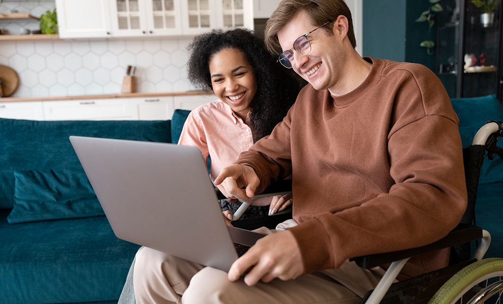 Inclusão digital. homem sentado em uma cadeira de rodas usando um laptop, enquanto uma mulher sentada ao lado dele também olha para a tela. Eles estão em uma sala com um sofá azul e uma cozinha ao fundo, que possui armários brancos e uma parede com azulejos hexagonais. O homem usa um moletom marrom e óculos, e a mulher veste uma camisa rosa.