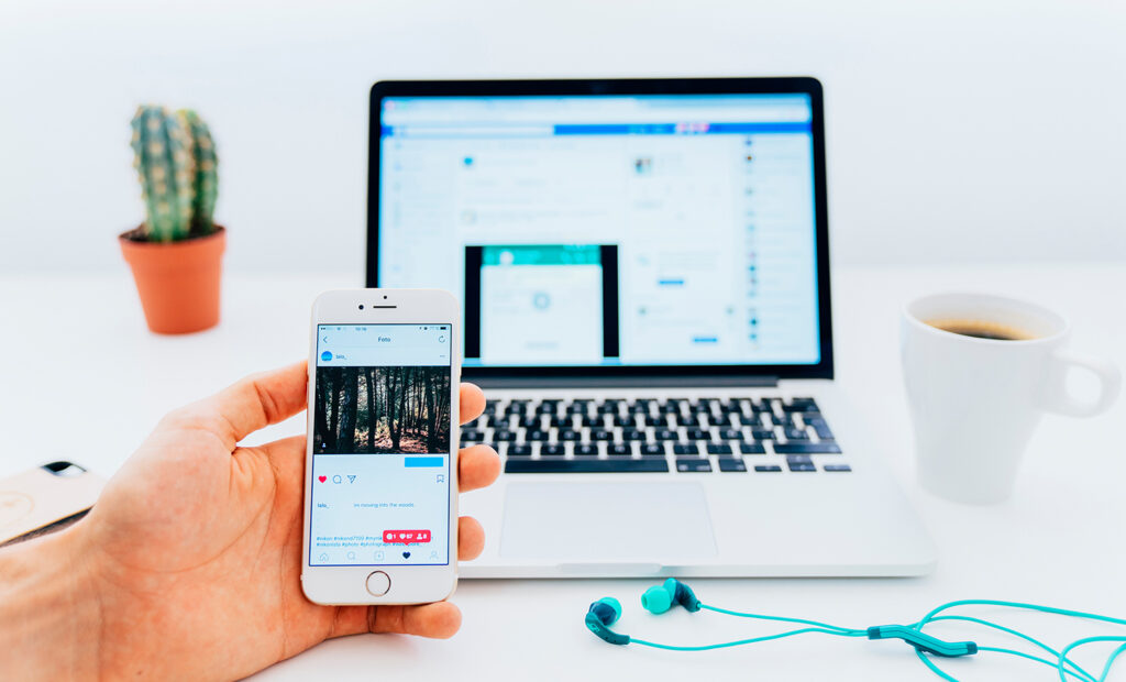 acessibilidade digital nas redes sociais e pessoas com deficiência: na imagem, há um laptop no fundo, mas em destaque está uma mão segurando um celular. Na tela do aparelho, aparece um post com um foto e as curtidas e comentários