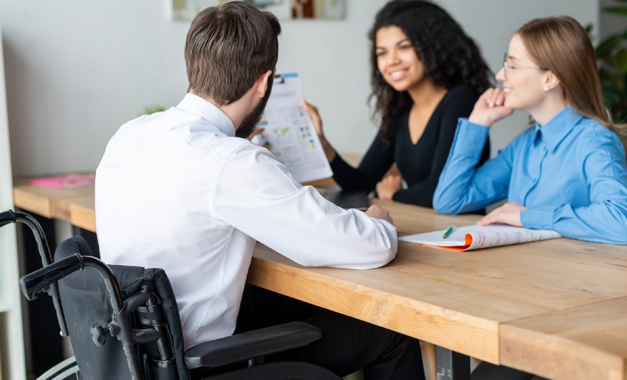 capacitismo nas empresas: duas mulheres estão sentadas em uma mesa de trabalho conversando com um homem cadeirante