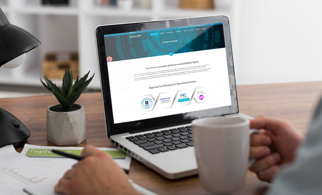 esg nas empresas e acessibilidade digital: na imagem aparece um laptop com a tela aberta. na tela, aparece uma das páginas da equalweb. aparece também o braço de uma pessoa apoiando na bancada, com uma mão segurando uma caneta e a outra uma xícara.