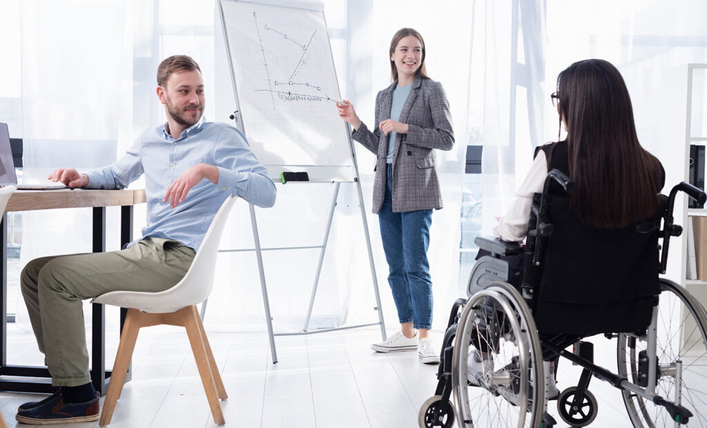 diversidade e inclusão: colegas de trabalho em um ambiente corporativo conversando e se olhando. Uma mulher está em pé ao lado de uma canaleta com uma caneta na mão, um cara está sentado com o braço apoiado na cadeira, e uma mulher, cadeirante, está de costas para a foto, olhando para ambos.