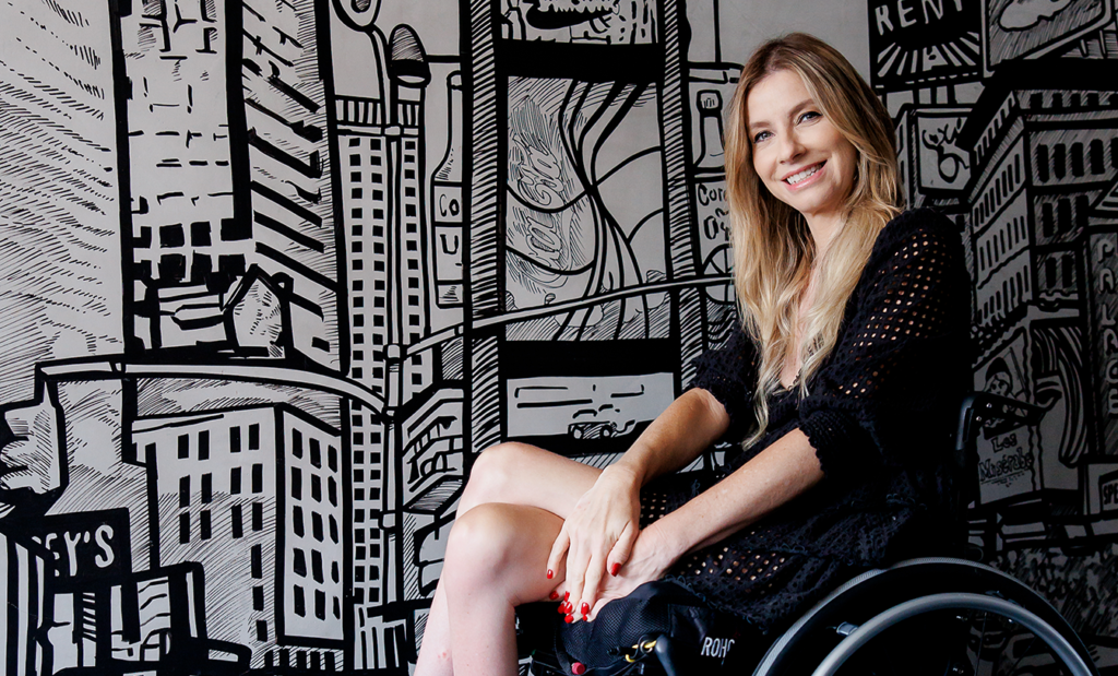 SAC. Imagem da Andrea nossa embaixadora sentada na cadeira de rodas com um vestido preto. Ao fundo temos um painel em preto e branco com a representação de alguns prédios.