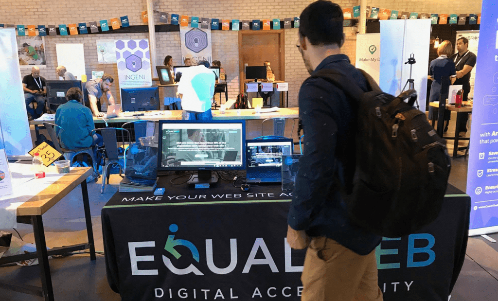 EqualWeb. Foto de uma feira aonde a EqualWeb tem um estande de apresentação. Na imagem aparece um rapaz de mochila nas costas e roupas sociais observando o stand da EqualWeb.