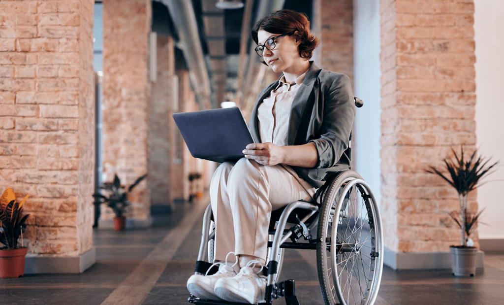 digital. Na imagem temos uma moça cadeirante de cabelos curtos, óculos e roupa social, usando um notebook.