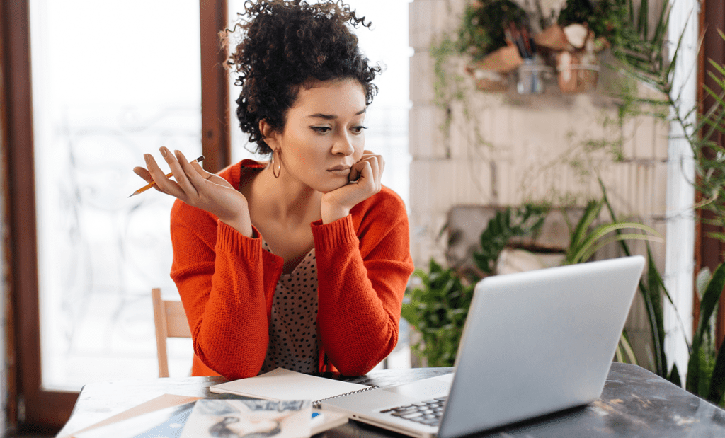 digital. Temos uma jovem brancar com cabelos cacheados e amarrados em um coque, na frente a tela do computador com uam feição pensativa. Ela usa um casaco na cor laranja.