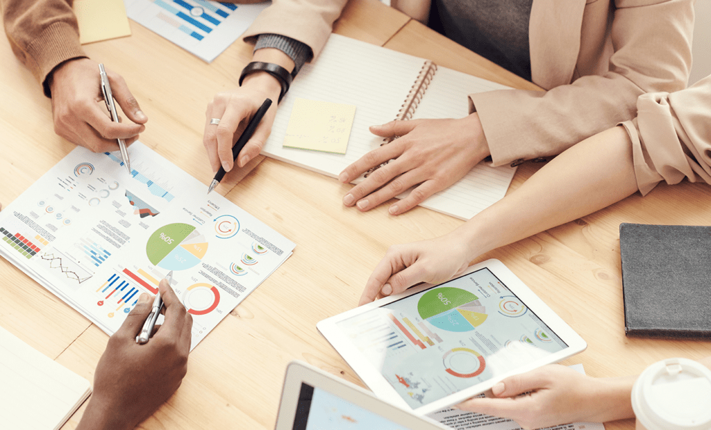 futuro. A imagem mostra uma mesa com alguns documentos, cadernos e tablets em cima. Nela também temos alguns mãos apontos para uma folha de relatório.