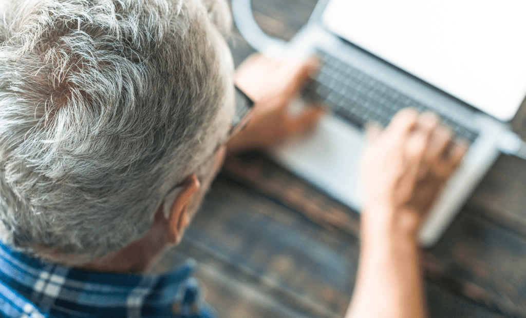 inclusão. Na imagem podemos observar um senhor de idade usando o computador . Ele tem cabelos grisalhoso e usa uma camisa xadrez, e também possui um aparelho auditivo.