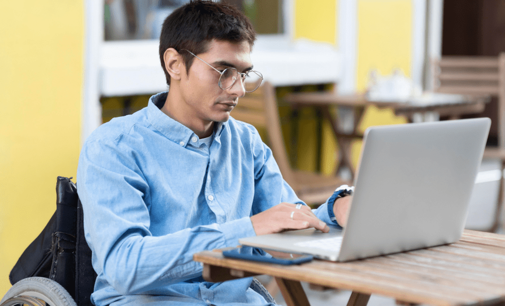 deficiência. Imagem de uma homem branco, cabelo preto usando óculos e cadeirante, sentado em um restaurante usando o notebook.