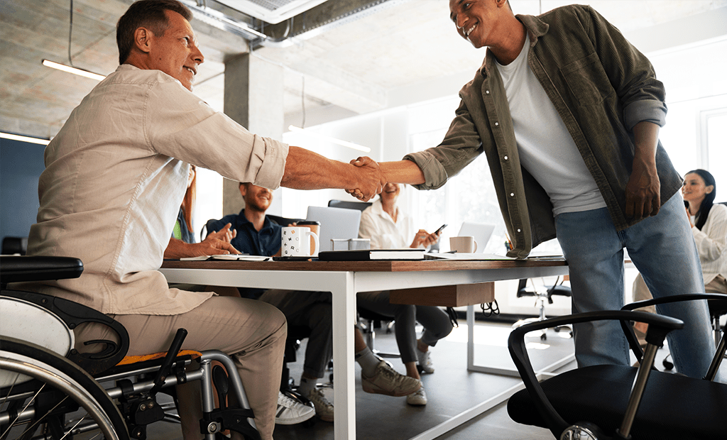 Empresa. A imagem representa uma sala de reunião com um grupo de pessoas. No angulo principal podemos observar dois homens apertando as mãos, um é cadeirante e o outro está em pé.