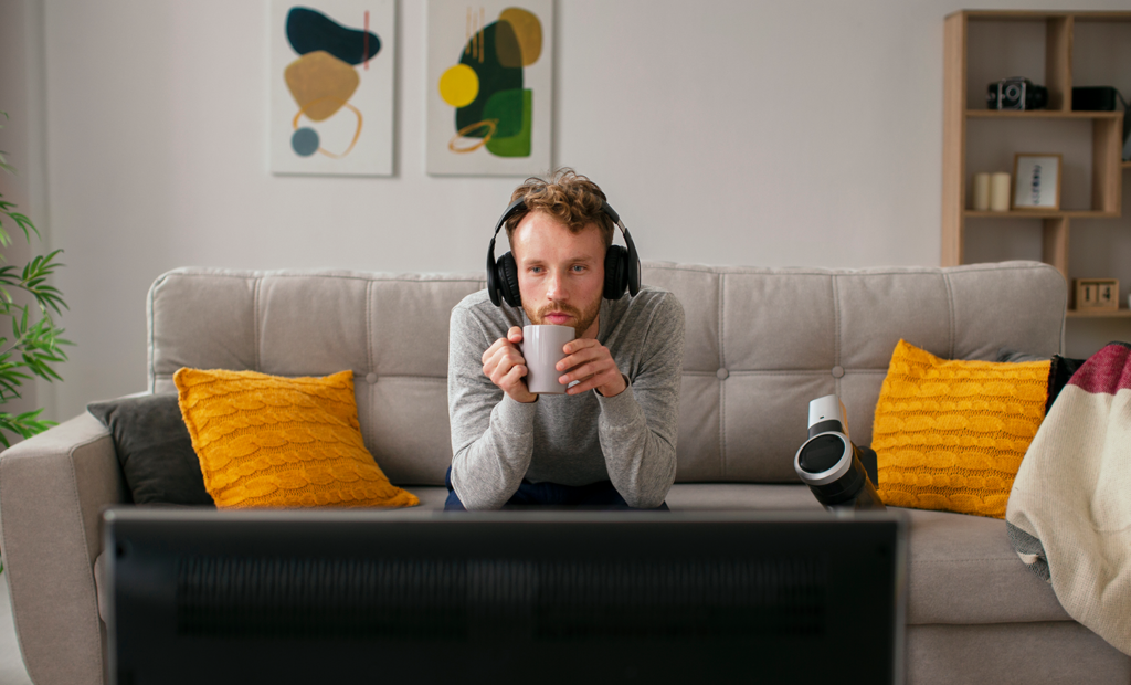 Campanha. Imagem de um homem branco, loiro. Ele usa uma casaco e um fone de ouvido. O homem está sentado na frente de uma televisão.