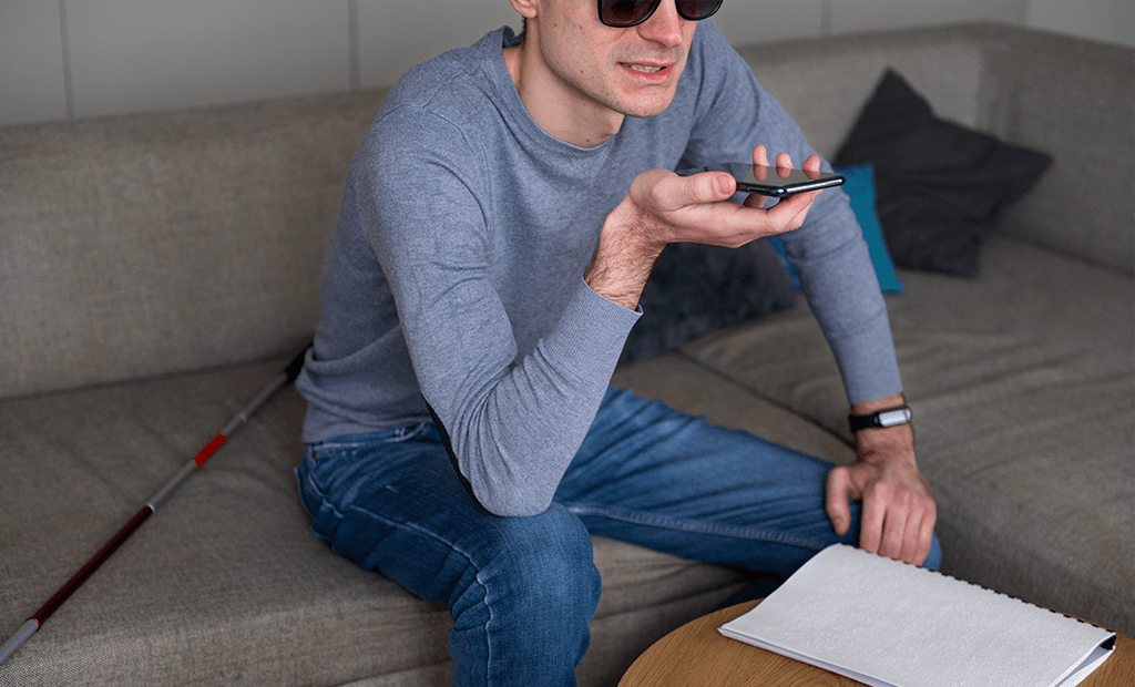 Design acessível. Imagem de um rapaz deficiente visual, sentado no sofá com a sua bengala do lado, falando ao telefone.