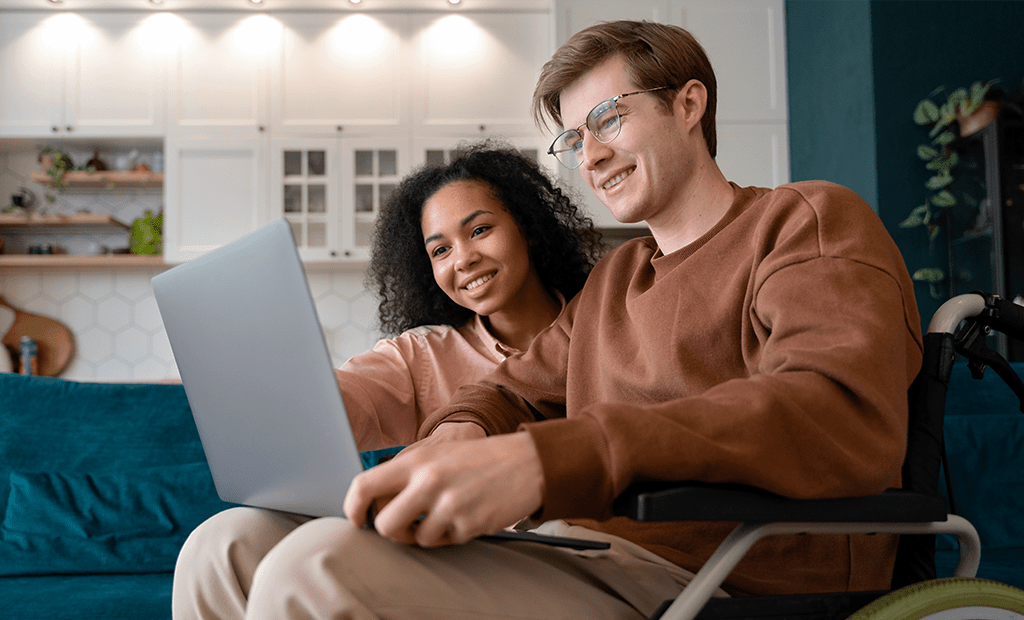 acessibilidade digital. Imagem de duas pessoas na frente do computador. Um deles é um rapaz brando de óculos, com casaco marrom, cadeirante e com um notebook no colo. Ao seu lado temos uma menina negra de cabelos pretos e casaco rosa apontando para o notebook.