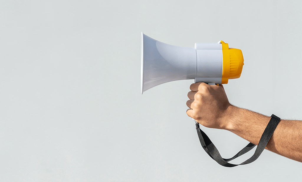 Comunicação. Imagem de uma mão masculina segurando um megafone. No fundo temos um parede branca.