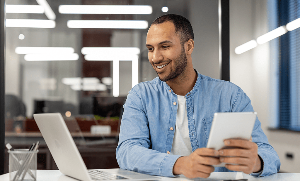 Epilepsia. Imagem de um homem negro aparentemente em um local de trabalho, usando jaqueta de frio e blusa branca. Ele está em uma bancada segurando um tablet com as duas mãos e olhando para o notebook que está em cima da mesa.