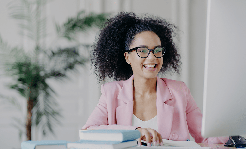 Acessível. Imagem de uma mulher negra de cabelos pretos e amarrados, vestindo um blaze rosa, sorrindo olhando para uma tela de computador.