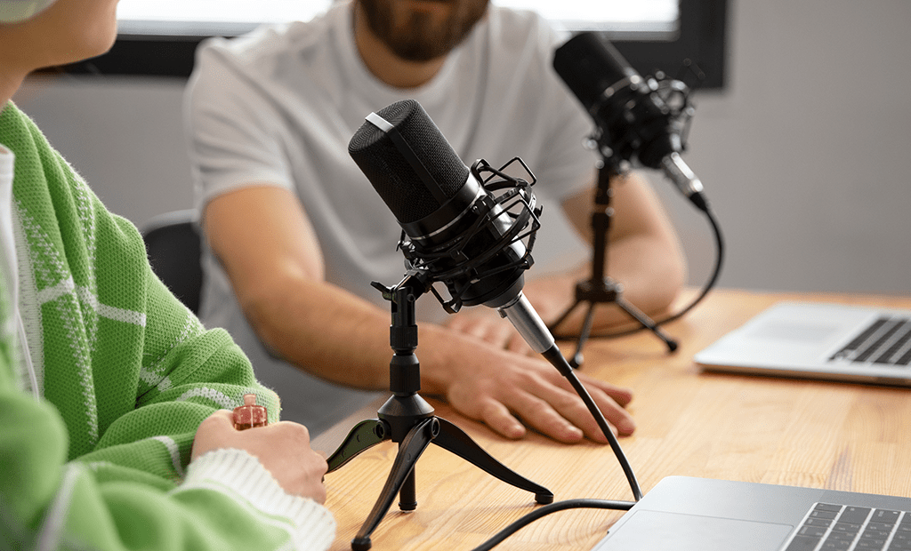 Acessibilidade Digital. Duas pessoas conversando em um podcast. Elas estão sentadas em uma mesa com microfones e notebooks. Uma pessoa veste um casaco verde e a outra uma camiseta branca.