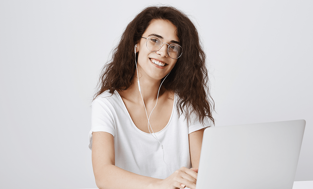 Acessibilidade Digital. Jovem sorridente, de cabelos cacheados e óculos, vestindo uma blusa branca e usando fones de ouvido enquanto trabalha em um laptop. O fundo é branco.