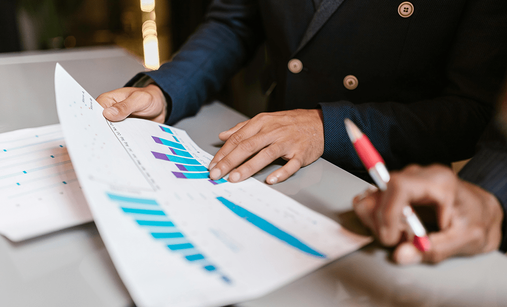 Acessibilidade Digital. Duas pessoas analisando gráficos impressos em um ambiente profissional. Uma delas aponta para os dados com o dedo, enquanto a outra segura uma caneta vermelha.