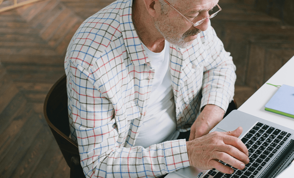 NBR 17225. Senhor idoso usando um notebook. Ele está vestindo uma camisa xadrez de cores claras e óculos. Ambiente interno, com um chão de madeira e uma mesa branca, onde há alguns cadernos ou blocos de notas ao lado do notebook.